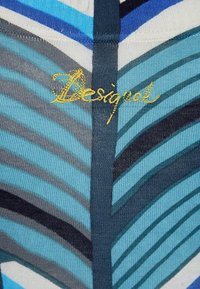 Gemusterter Stoff in verschiedenen Blau- und Grautönen mit einem Blättermuster. Gesticktes "Desigual"-Logo in goldfarbener Faden.