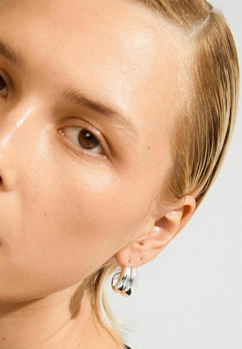 Detailný záber na tvár osoby so svetlou pokožkou, hnedými očami, hladko stiahnutými blond vlasmi a strieborným dvojitým kruhovým náušníkom v pravom uchu.