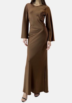 Kvinde iført en lang brun satin kjole med lange flagrende ærmer og sorte åbne tå-hæle, stående mod en hvid baggrund.