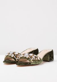 Sandaalit, joissa on avonainen kärki, vihreää samettia ja paksu korko, varustettuna jalokivillä koristellulla hihnalla, jossa on eri kokoisia kirkas- ja pastellisävyisiä kiviä.