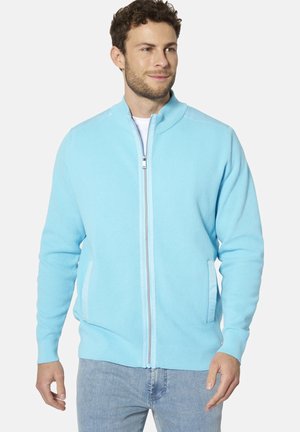 Mann trägt hellblaue, strukturierte Reißverschlussjacke und hell gewaschene Jeans, schaut leicht nach rechts mit neutralem Gesichtsausdruck.