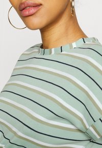 T-shirt às riscas em verde claro com linhas horizontais em bege, branco e azul-marinho. Apresenta um decote redondo e um tecido macio e texturizado.