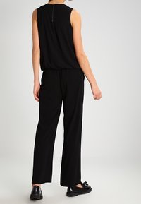 Femme portant un haut noir sans manches et un pantalon noir ample, se tenant de dos face à l'appareil photo sur un fond clair.