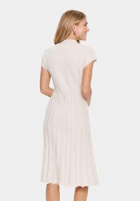 Knit dress in light beige with a fitted bodice and pleated skirt. Features short sleeves and a soft, textured fabric with horizontal stripes.