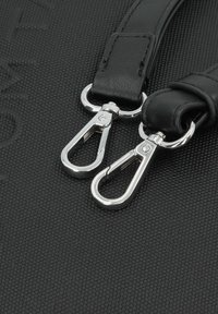 Des sangles en cuir noir avec des clips en métal argenté, présentant une surface texturée, fixées à un fond en tissu sombre.