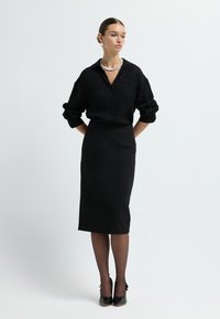 Schwarze Bluse mit Knopfverschluss und gemusterter Textur, kombiniert mit einem figurbetonten schwarzen Midi-Rock, transparenten schwarzen Strumpfhosen und schwarzen Absätzen mit Schleifen.