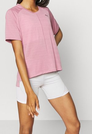 Mujer con una camiseta rosa suelta de Puma y pantalones ciclistas blancos ajustados, posando contra un fondo claro y sencillo.