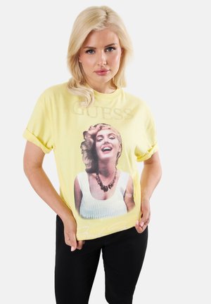 Femme blonde portant un t-shirt jaune aux manches retroussées avec un motif d’une femme souriante, associée à un pantalon noir sur fond blanc.