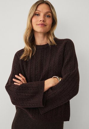 Vrouw met blond haar die een donkerbruine kabelgebreide trui met wijde mouwen en een gouden armband draagt, staand tegen een effen lichte achtergrond.