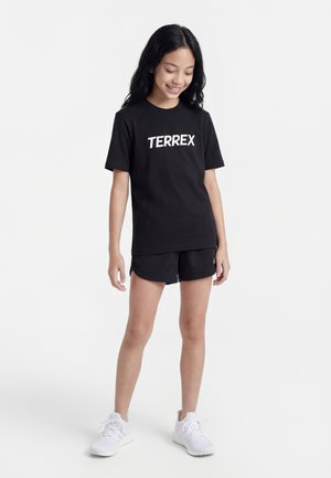Mladé dievča s dlhými tmavými vlasmi, ktoré má na sebe čierne tričko TERREX, čierne šortky a biele tenisky, stojí na bielom pozadí.
