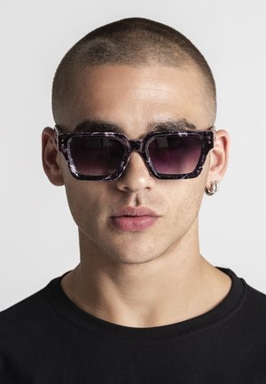 Man with buzz cut wearing large, square, dark purple tortoiseshell sunglasses and silver hoop earrings, dressed in a black shirt, against a plain background.