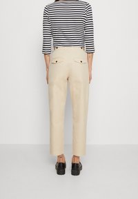 Beige kalhoty se střihem do rovna, obsahující dvě zadní kapsy a knoflíkové akcenty. Model má na sobě modré pruhované triko.