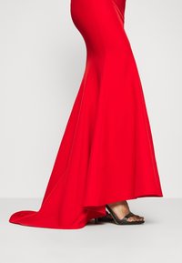 Robe sirène rouge jusqu'au sol avec un léger traîne, portée avec des sandales à talons noires à bout ouvert.