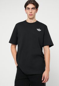 Černé bavlněné tričko s krátkými rukávy, které má na levé hrudi malé bílé logo Adidas. Jednoduchý, klasický design; volný střih.