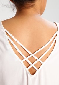 Bílý top s V-výstřihem na zádech a zkříženými bílými popruhy, hladká textura a volný střih, který zvýrazňuje odhalenou pleť pod ním.