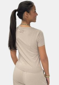 Camiseta ajustada de color beige claro, confeccionada en tela suave, con cuello redondo y mangas cortas. Logo de EA7 impreso cerca del escote.