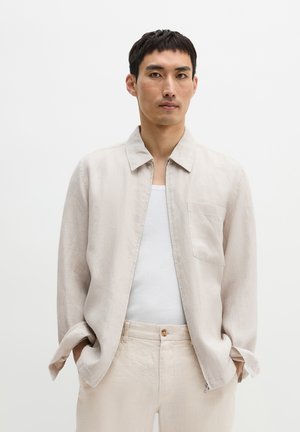 Mężczyzna w jasnobeżowej kurtce na białym podkoszulku, z rękami w kieszeniach, na jednolitym białym tle.