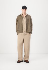 Jachetă oversize în carouri maro, cu buzunare frontale, cămașă bej cu nasturi purtată dedesubt, pantaloni bej și pantofi maro. Căciulă neagră purtată.
