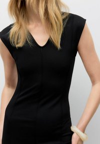 Robe noire ajustée avec un décolleté en V et des manches courtes, dotée de détails de couture. Portée avec un bracelet translucide au poignet.