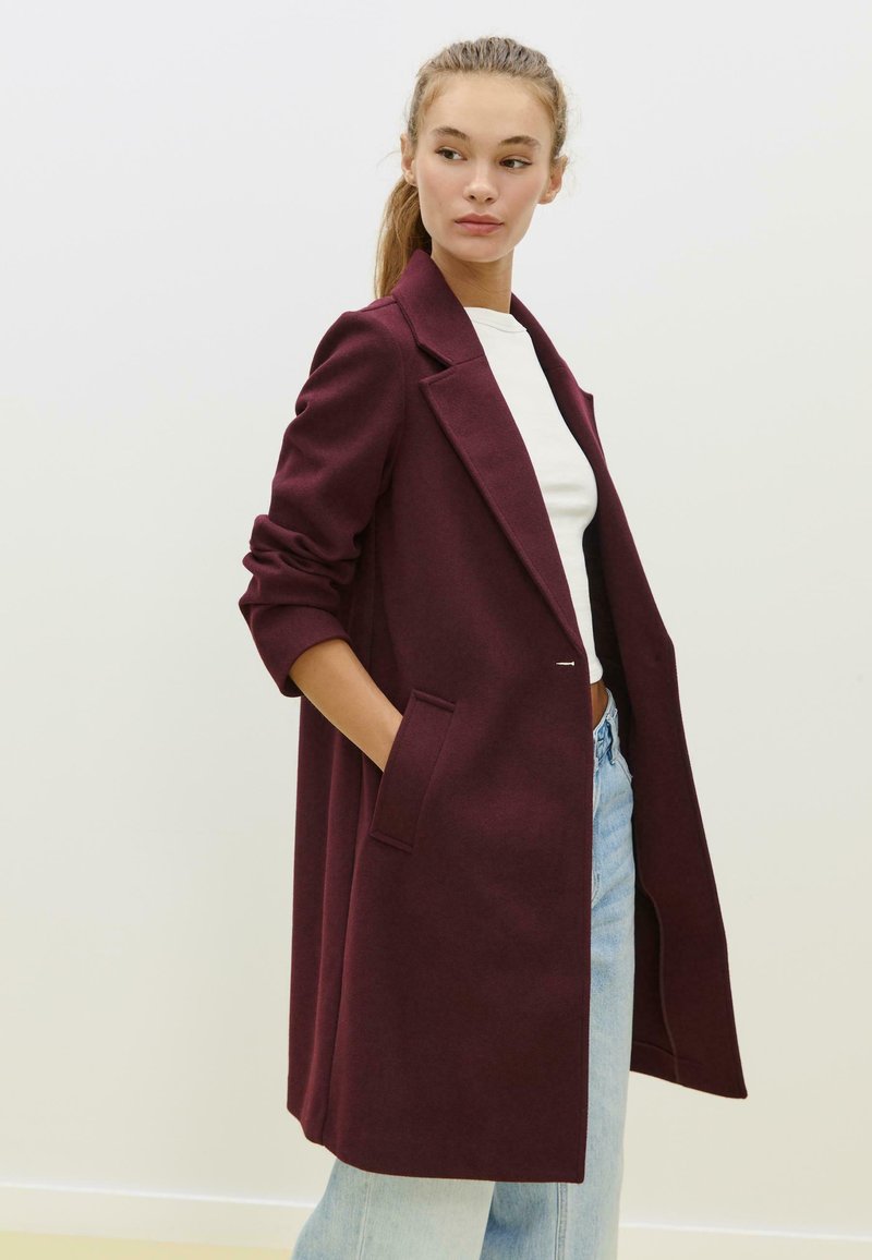 Manteau en laine bordeaux avec un col cranté, des manches longues et des poches latérales, présentant une silhouette décontractée et une texture douce.