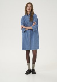 Denim shirt dress, oversized fit, light blue color, buttons down the front, short sleeves, chest pocket, worn with dark socks and shoes.