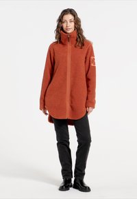Fleecejakke i brændt orange med høj krave, fuld lynlås, sidelommer og tekstureret overflade. Sammen med sorte bukser og støvler.