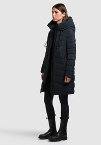 Töltött hosszú kabát sötét navy kék színben, magas gallérral, zipzáros elejével, átlós steppelt mintával és oldalsó cipzáras zsebekkel. Fekete leggings-szel és csizmával viselve.