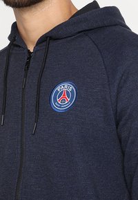 Sudadera con capucha de cremallera en color azul marino con una textura suave, que presenta un logo circular del Paris Saint-Germain en el área del pecho izquierdo y detalles de cordones.