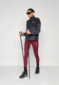 Fekete és szürke félcipzáras dzseki, piros leggings, fekete túracipő. Túrabotokkal a kezében. Napszemüveg és márkajelzés látható a dzsekinken.