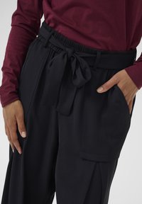 Pantalons larges noirs avec une taille à nouer, dotés de poches latérales et d'un tissu lisse et texturé. Associés à un haut à manches longues bordeaux.