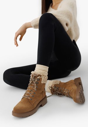 Botine maro din suede cu o margine de blană texturată, cu un design cu ochiuri din metal, cu șireturi și talpă din cauciuc, asortate cu șosete din tricot crem.