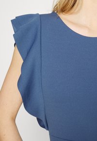 Gros plan d'une robe bleue avec des manches courtes à volants superposés portée par une personne à la peau claire aux cheveux blonds.