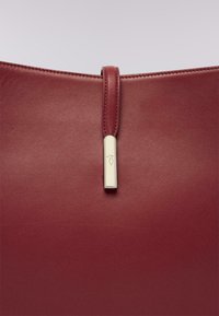 Borsa in pelle bordeaux con una texture liscia, dotata di una tracolla centrale a loop e un accento in metallo dorato a contrasto.