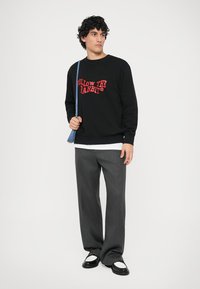 Sweat-shirt noir avec le texte rouge "SUIVEZ LE LAPIN", pantalon gris, chaussures noir et blanc, et un sac à épaule bleu clair.