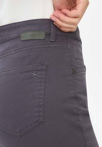 Grå denimbukser med en let elastisk tekstur, med et grønt lædermærke, tonede syninger og et lille broderet logo nær lommen.