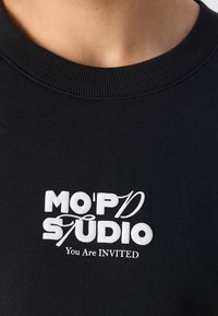 Pull noir à col rond avec texte blanc "MO'P'D STUDIO" et texte plus petit "Vous êtes INVITÉ" centré sur la poitrine.