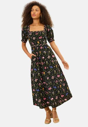 Femme aux cheveux bouclés portant une robe midi noire à fleurs avec des manches bouffantes et un décolleté carré, debout avec une main dans la poche et des sandales dorées.