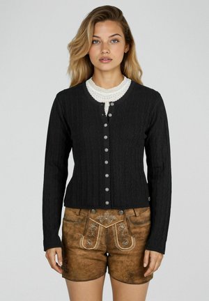 Femme portant un cardigan noir à boutons en maille torsadée par-dessus un haut en dentelle blanche et un short en cuir brun brodé, debout devant un fond uni.