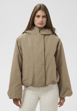 Ung kvinde med langt brunt hår iført en stor beige jakke og hvide teksturerede bukser, står mod en ensfarvet lys baggrund.