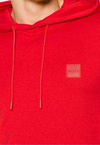 Sudadera roja con capucha ajustable y un logo cuadrado con "BOSS" en relieve, fabricada con un tejido suave y un acabado texturizado.