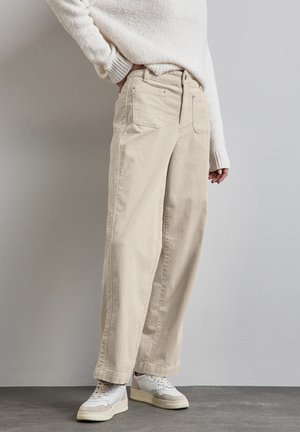 Pantalons en velours côtelé beige à jambes larges avec poches latérales et arrière, associés à un pull blanc texturé et des baskets blanches avec des accents gris.