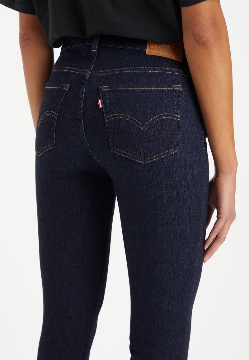 Pantalones vaqueros de mezclilla azul oscuro con cintura alta, costuras visibles y dos bolsillos traseros con un diseño curvado característico. Parche de logo de cuero en la cintura.