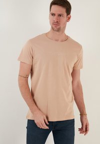 Camiseta de manga corta en un suave color beige claro, con un cuello redondo y un sutil diseño de texto en relieve en la parte delantera, confeccionada en suave tejido de algodón.