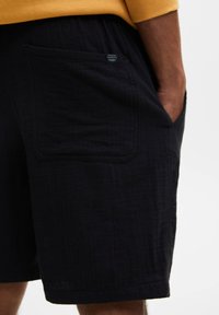 Pantalones cortos negros con un bolsillo lateral visible y una pequeña etiqueta de tela, llevados por una persona con una mano metida en el bolsillo, sobre un fondo liso.