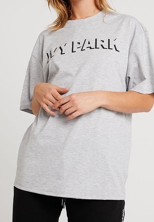 Nainen pukeutuneena vaaleanharmaaseen ylipitkään t-paitaan, jossa lukee "IVY PARK", ja mustiin housuihin, kädet rentona edessä yhteen kiedottuina.