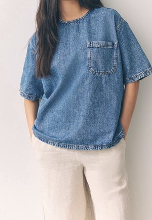 Femme portant une chemise ample en denim bleu à manches courtes avec une poche avant et un pantalon en lin crème, mains dans les poches.