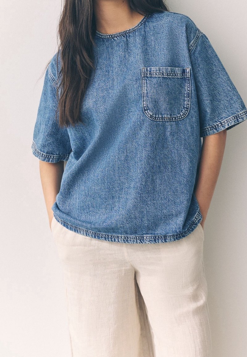 Femme portant une chemise ample en denim bleu à manches courtes avec une poche avant et un pantalon en lin crème, mains dans les poches.