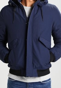Mann trägt eine marineblaue, mit Kapuze versehene Winterjacke mit Fronttaschen und schwarzen Bündchen, kombiniert mit einem grauen Hemd und blauen Jeans.