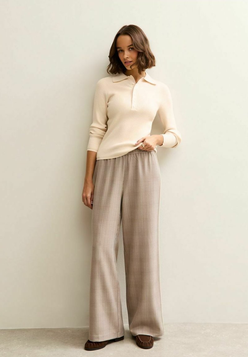 Maglione a coste beige con collo, abbinato a pantaloni a gamba larga a quadri in tonalità marrone chiaro. Scarpe slip-on marroni completano il look.