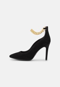 Scarpa nera in suede con tacco alto, punta affusolata, una decorazione in catena dorata alla caviglia e una superficie liscia.
